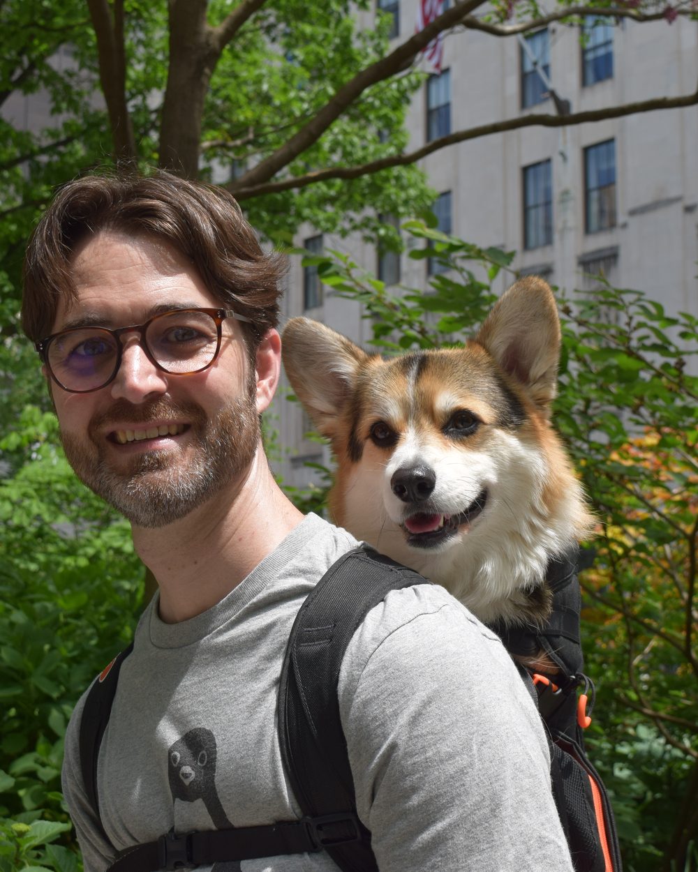 Aydrian smiling next to Atticus the corgi in a chest carrier in a Manhattan park.